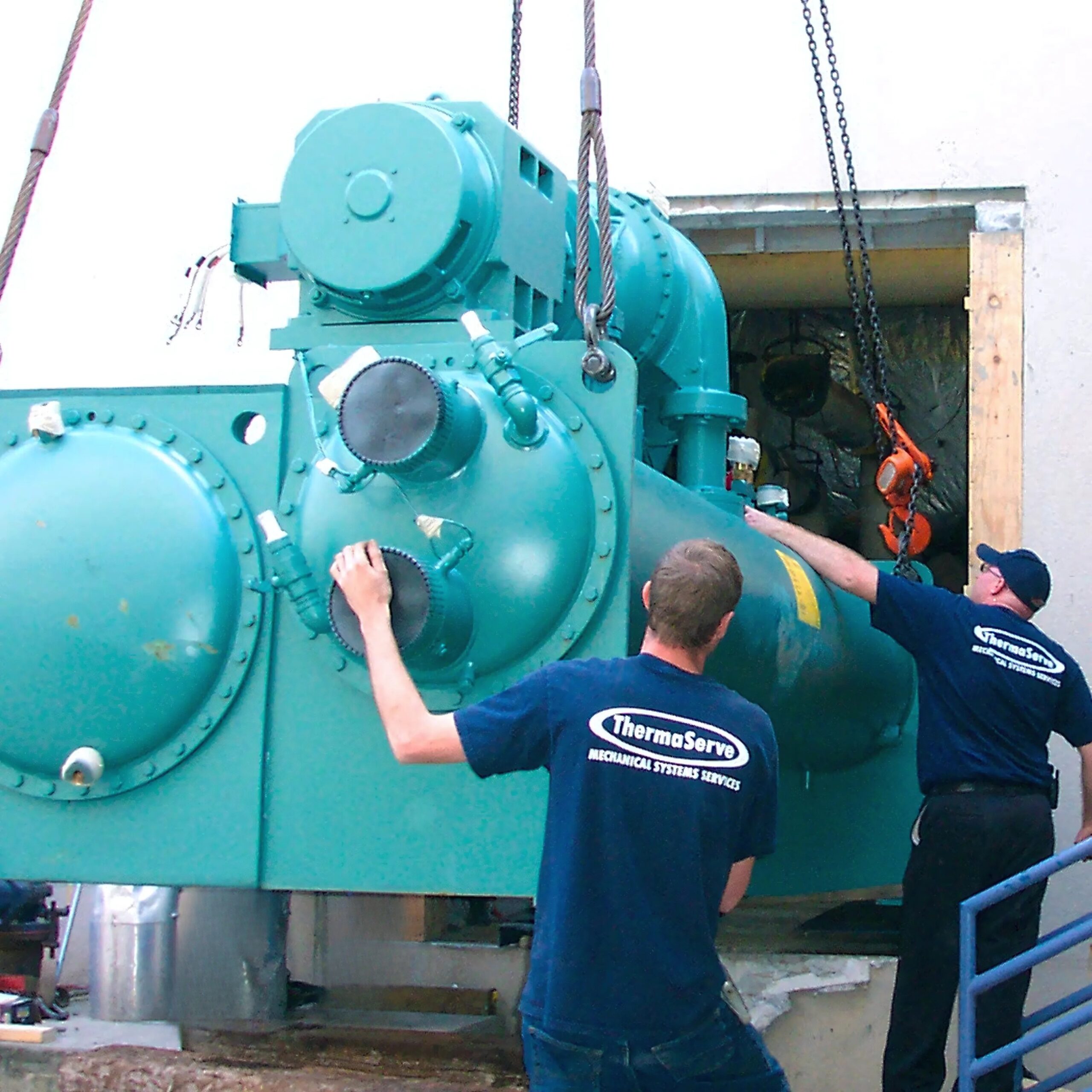 ThermaServe technicians guide a large industrial chiller into a building using crane hoists.