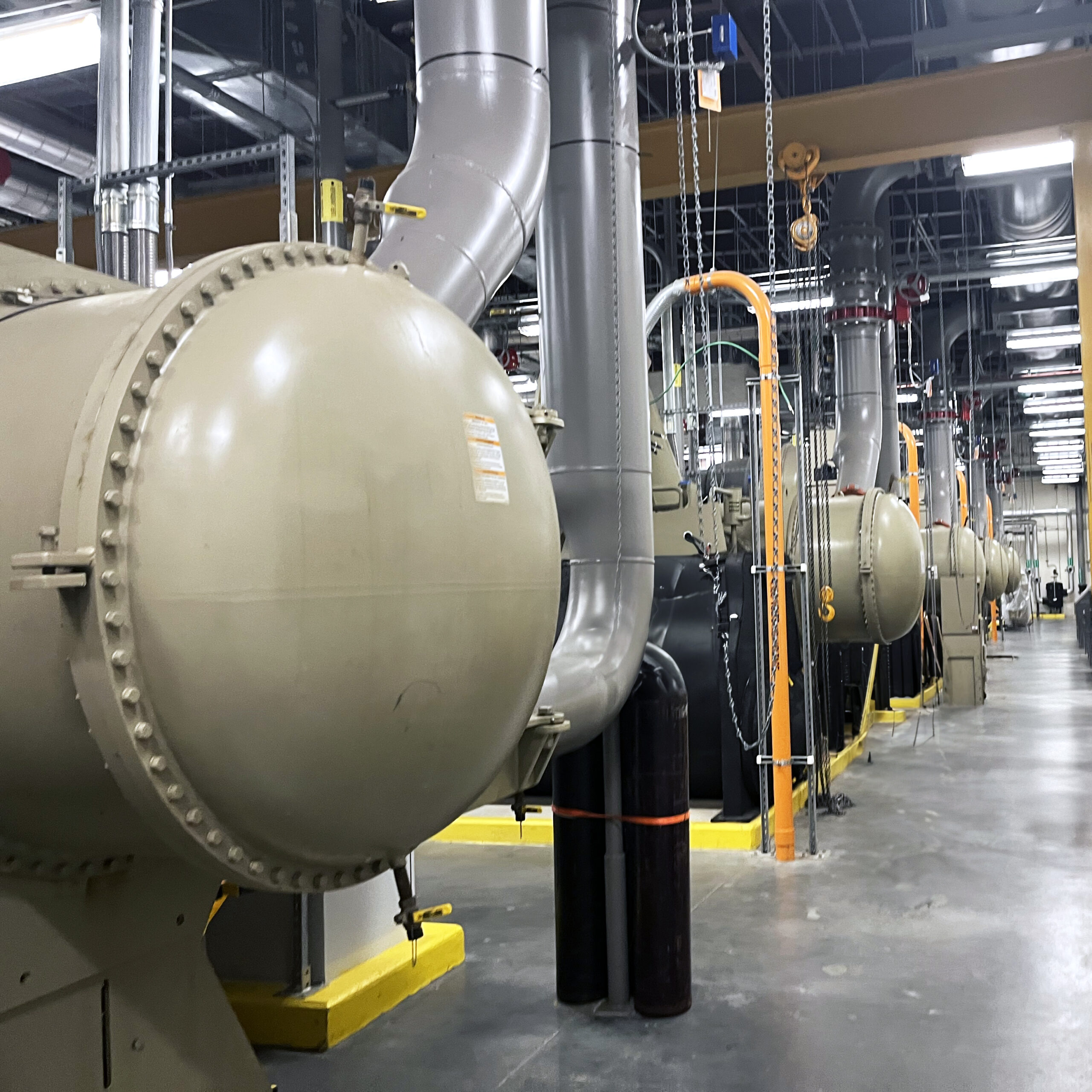 Row of large industrial Trane chillers with overhead piping in a commercial mechanical room.